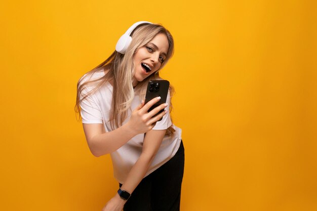 A girl sings to music in headphones with a connected phone on a yellow background