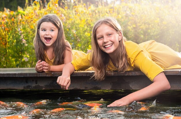 Photo girl sisters lie on the bridge and feed the fish in the park outdoors