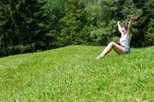 Una ragazza si siede su uno sfondo di montagne sul prato verde e gode del sole.