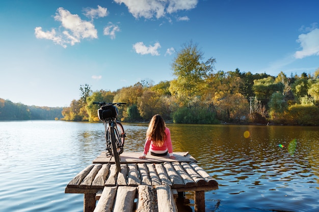 Photo girl sits on the bank of the river. bike on the river with a bag on the trunk