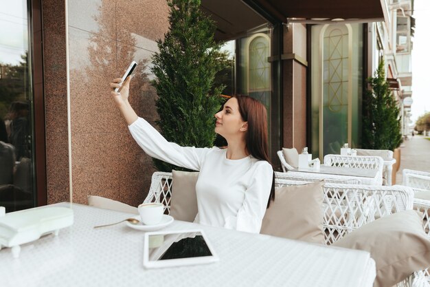 The girl sits in a cafe using technology. the girl looks at a teleon in a cafe at a table.