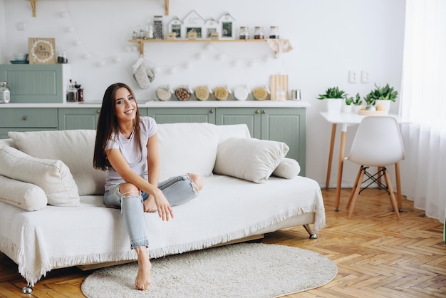 The girl sits on the couch with one leg bent and looks at the camera