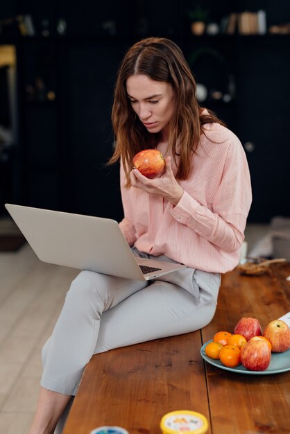 Girl sits on desk with laptop and eats apple.