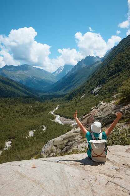 A girl sits on the edge of a cliff and looks into the distance in the distance there is a river and a gorge a good mood clouds and a mountain landscape