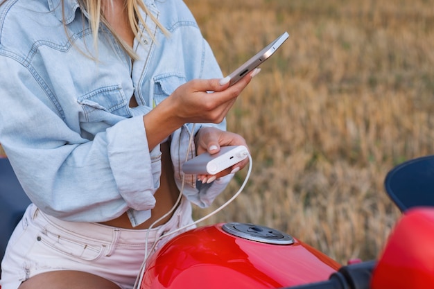 女の子がバイクに座って、白い画面のスマートフォンのモックアップを持っています。パワーバンクは、自然を背景に携帯電話を充電します。