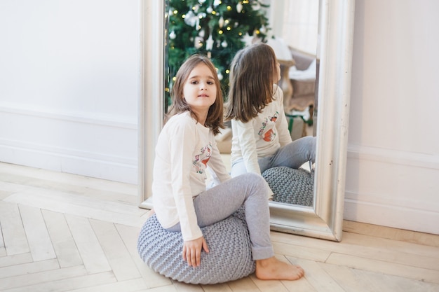 The girl sits near the mirror, the mirror reflects the Christmas tree and lights