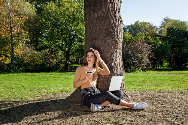 A girl sits near a tree in a park with a cup of coffee and works on a laptop Freelance w