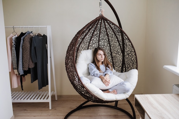 The girl sits in pajamas in a cocoon chair. Stylish interior of the room. The girl is sad.