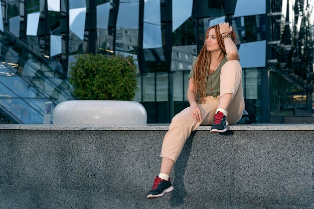 Girl sits on the parapet hanging her leg down. Against the background of an office building.Non-standard office employee