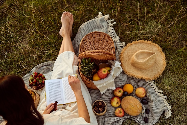 The girl sits on a plaid in the open air and reads a book.