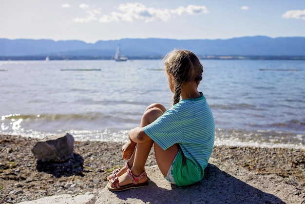 女の子はジュネーブ湖の岸壁に座って、水と山の休暇と夏の旅行を見ています