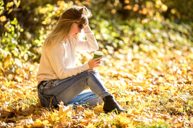 La ragazza si siede e si rilassa per terra nella foresta autunnale con il caffè