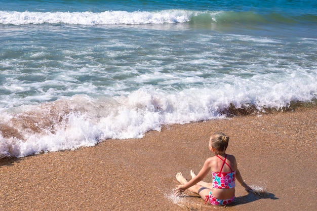 The girl sits on the sand on the seashore with waves Children39s summer sea vacation