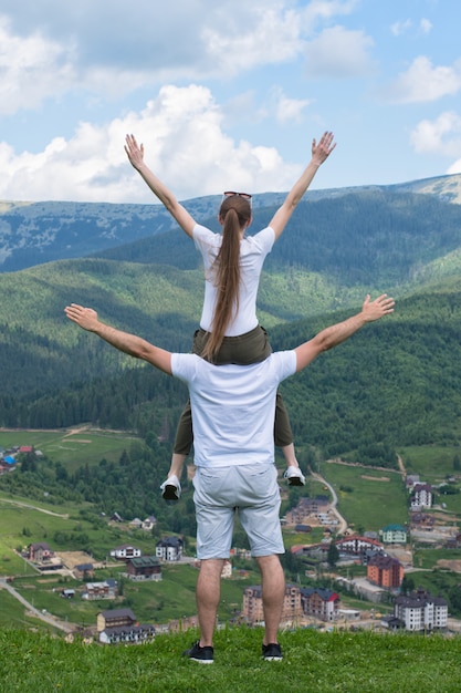 Girl sits on the shoulders of the guy with hands raised. Mountains on the background. Back view