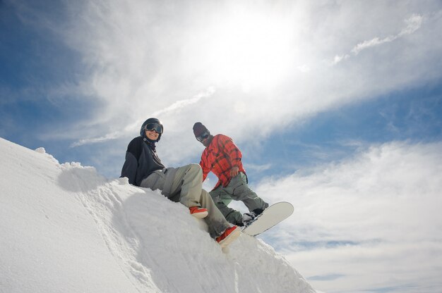 Girl sits on the snow on the hillside , and the guy is getting ready to go down on the snowboard