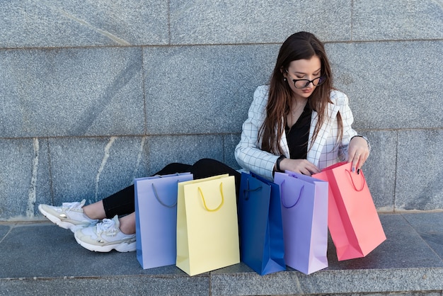 Girl sits on the stairs and considers purchases in colored bags. After successful shopping.