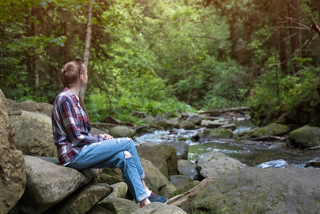 Girl sits on stone on the mountain river and forest. Nature walks