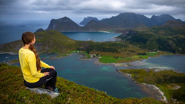 소녀는 로포텐 섬, 노르웨이 하이킹의 파노라마를 즐기는 오퍼소이카멘 꼭대기에 앉아 있습니다.