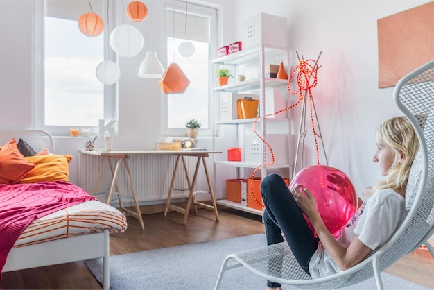 A girl sits in a white chair in a room with a pink balloon.