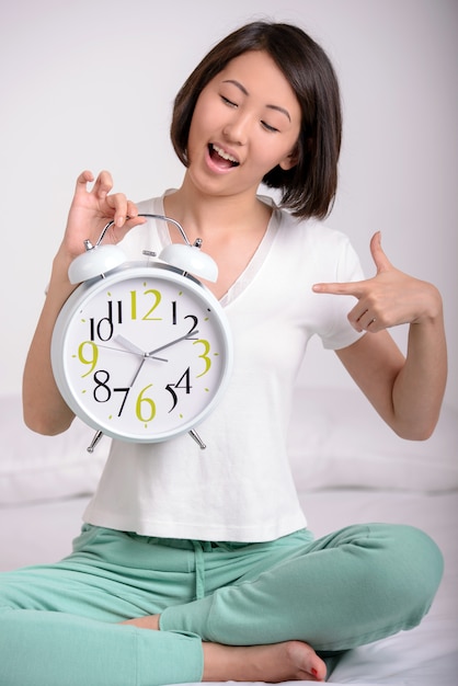 Girl sitting on the bed, holding big alarm clock.