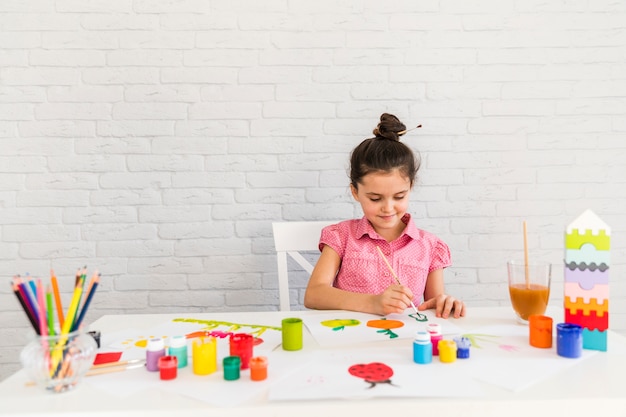 Una ragazza che si siede sulla sedia pittura su carta bianca con bottiglia di vernice colorata e matite colorate sul tavolo