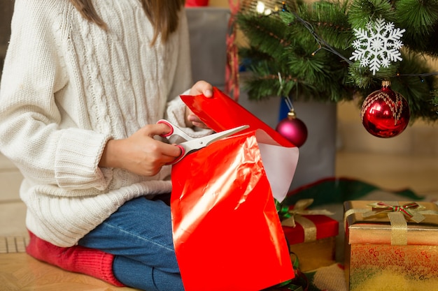 Girl sitting under Christmas tree and cutting red paper with scissors for decorating presents