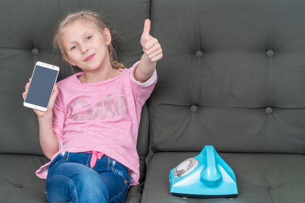 girl sitting on the couch next to a retro phone shows the screen of her smartphone, lifting a thumbs up