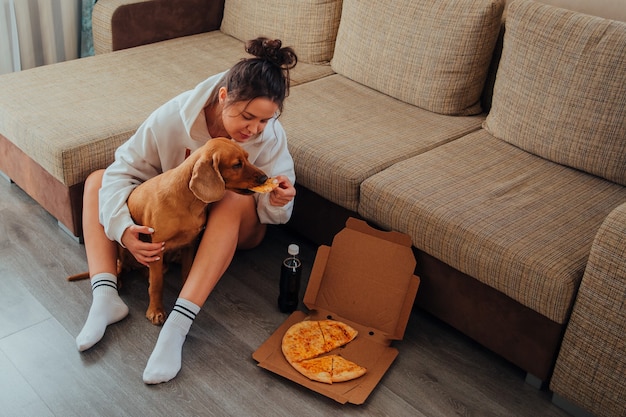 Girl sitting on the floor and feeding Cocker Spaniel pizza, leaning on the sofa