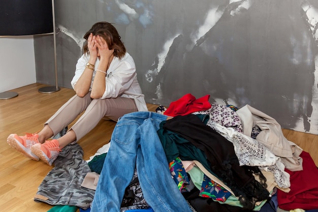 A girl sitting on the floor sorts out her wardrobe a mountain of things in the room against