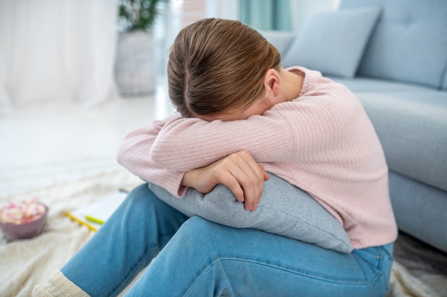 Girl sitting on the floor with her head down on her hands, which are on the pillow, hiding her face.