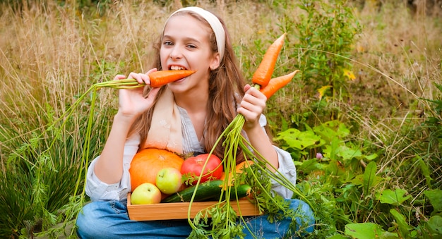 Фото Девушка сидит на траве и ест свежую морковь, свежие яблоки, огурцы, тыкву в корзине