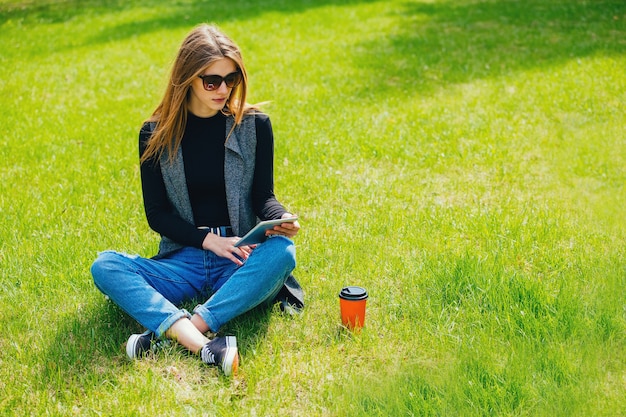 写真 通りでコーヒーとタブレットを持って草の上に座っている女の子。