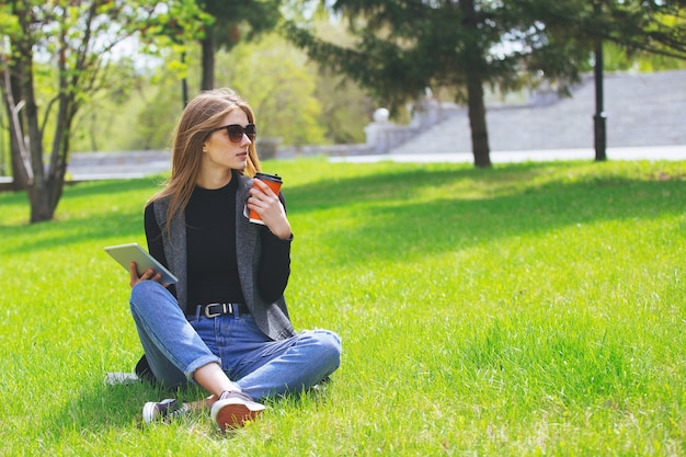 写真 通りでコーヒーとタブレットを持って草の上に座っている女の子。