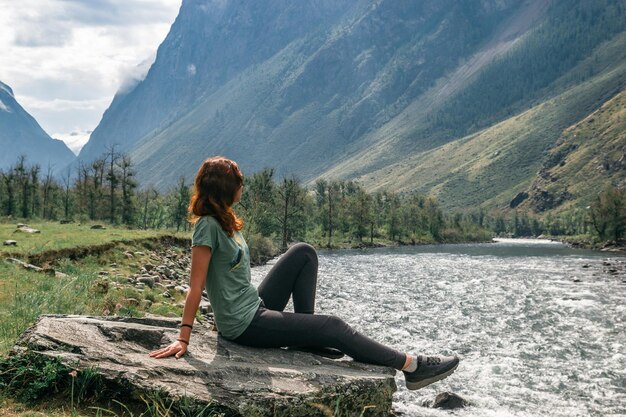 Girl sitting on a stone on the river bank against the backdrop of mountain slopes