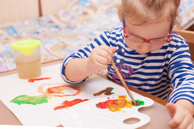 Photo girl sitting at the table and draws paints on white paper