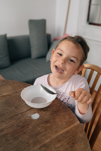 A girl sitting at a table with a spoon in her hand and a grey couch behind her.