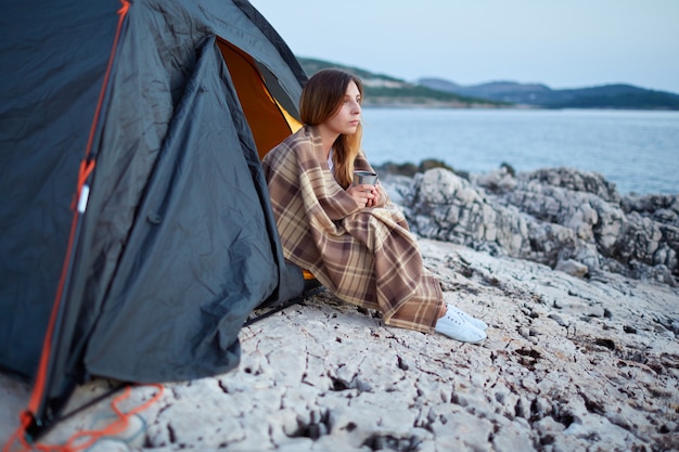 Photo girl sitting under tents, wrapped in plaid, holding cup of fragrant tea.