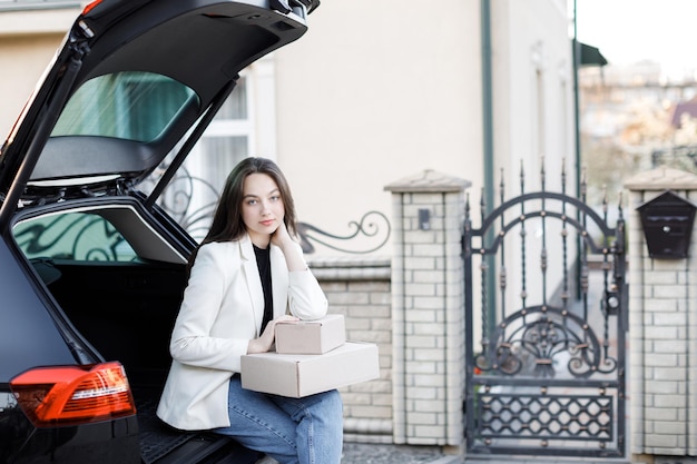 車のトランクに座って箱を手に持って、カメラを見て笑っている女の子オンラインで商品を購入して家に届けるというコンセプト