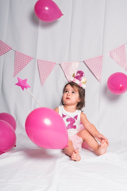 Una ragazza seduta con una bacchetta magica e palloncini rosa su fondo bianco.