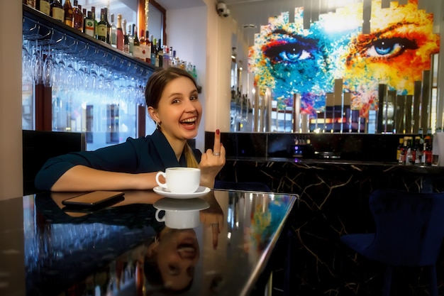 Girl of Slavic appearance in bar, cafe or restaurant with white Cup. Female shows emotion. Young charming woman is sitting at bar and drinking coffee or tea in her free evening time