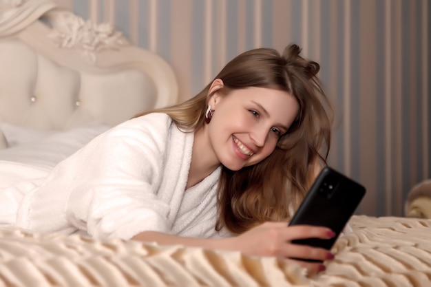 Girl of Slavic appearance in white coat with phone on bed in hotel room