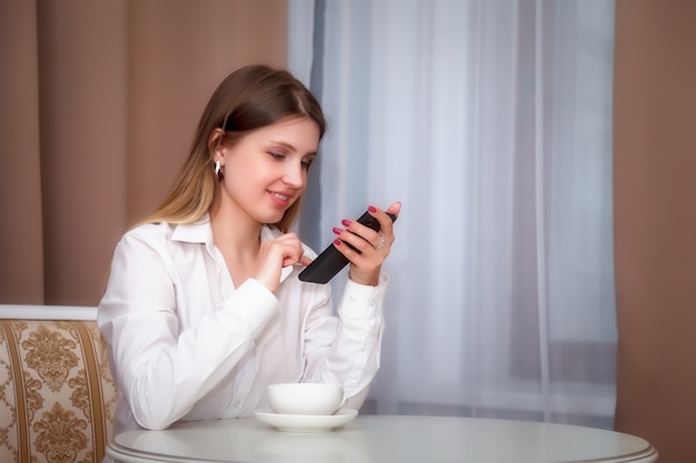 Girl of Slavic appearance with phone and white Cup is sitting at table