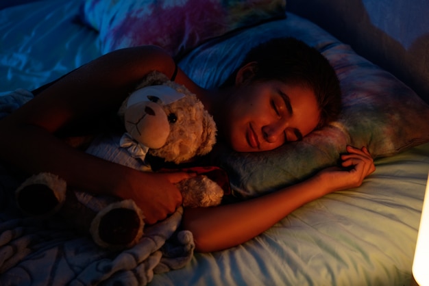 柔らかいおもちゃ、夜の光からの光が付いているベッドで寝ている女の子
