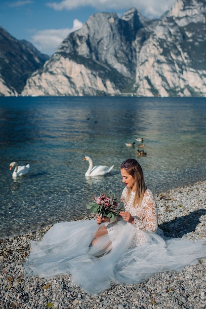 Una ragazza con un elegante abito bianco è seduta sull'argine del lago di garda, mentre una donna è fotografata sullo sfondo di una montagna e di un lago in italia.torbole.