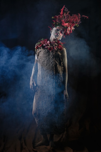A girl smeared with clay in a cemented dress. The model has a headdress made of flowers. Smoke from behind