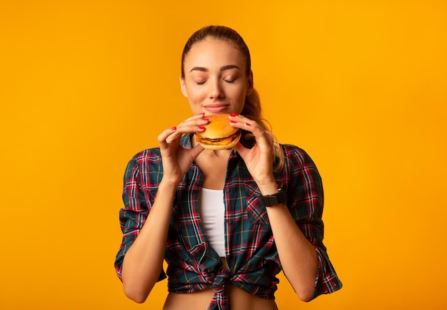 黄色のスタジオの背景の上に立ってハンバーガーの匂いを嗅ぐ女の子