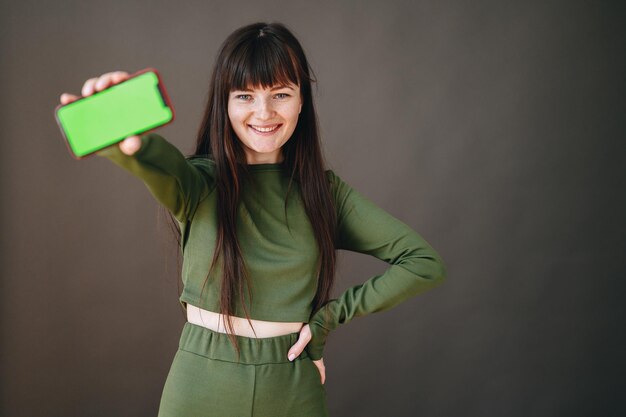 Girl smiles broadly with her hand outstretched in which phone with green screen and hand on her hip