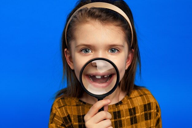 Girl smiling brightly through magnifying glass isolated on blue wall