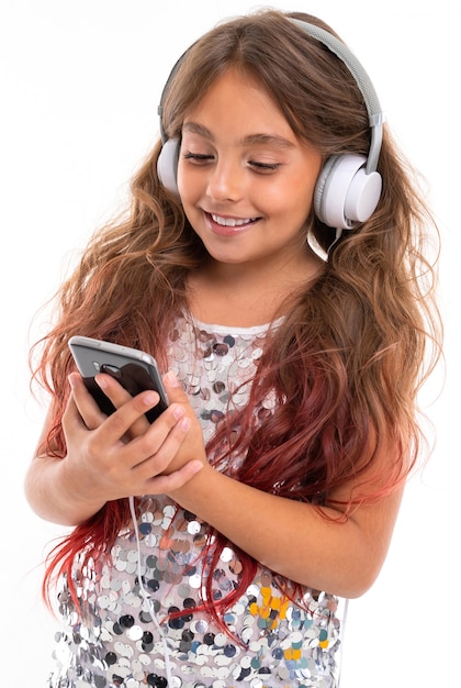 Girl in sparkling dress, with big white earphones listening to music and staring at screen of black smartphone isolated