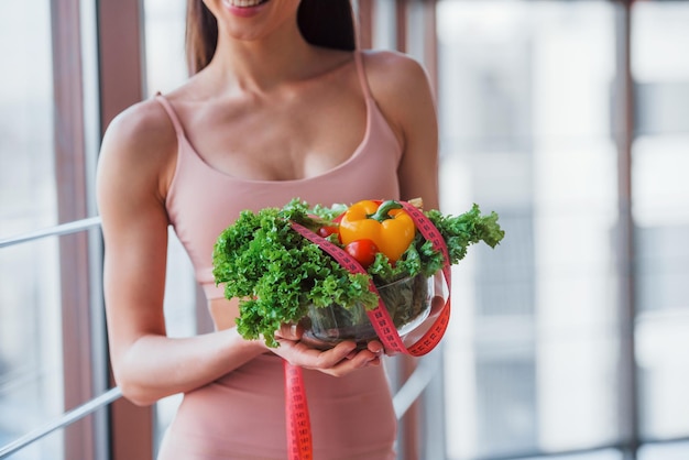 Girl in sportive wear and with healthy food in hands stands indoors near window.